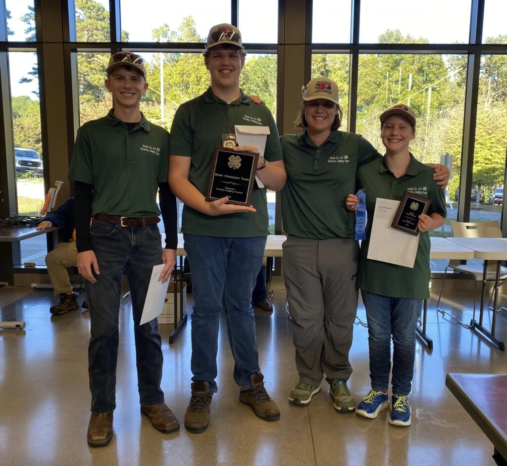 Four 4-H'ers in green polo shirts smile as they hold up ribbons and award plaques